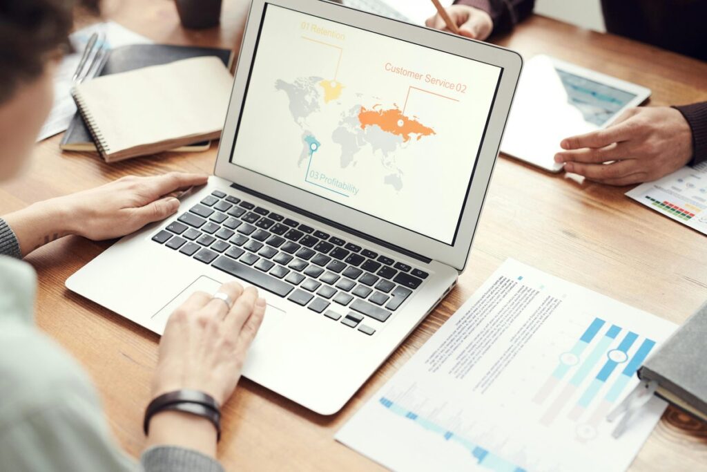 Laptop showing a slide with a world map and three labeled points, with printed charts, notebook and tablet on a desk