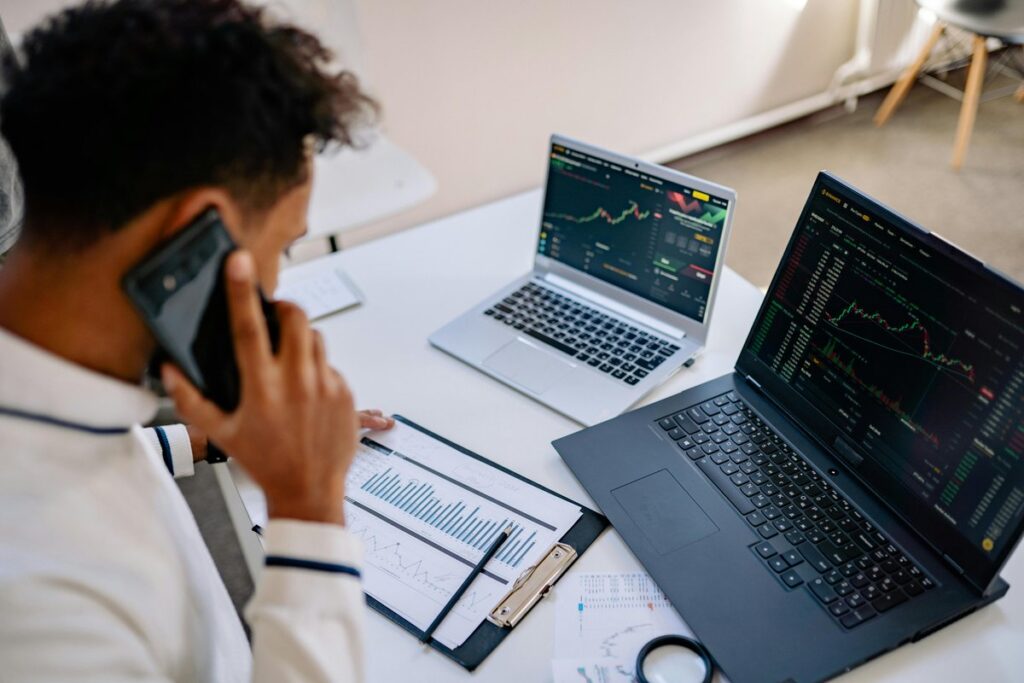 Salesperson on the phone reviewing printed charts and two laptops before a sales meeting